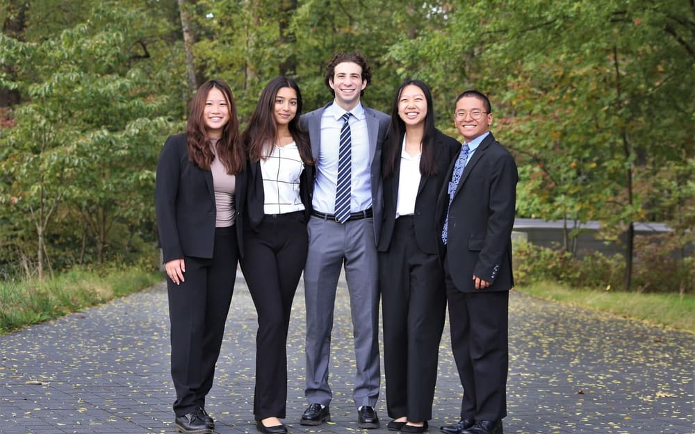 Five SCG members in business attire standing together on a paved path in front of green trees.