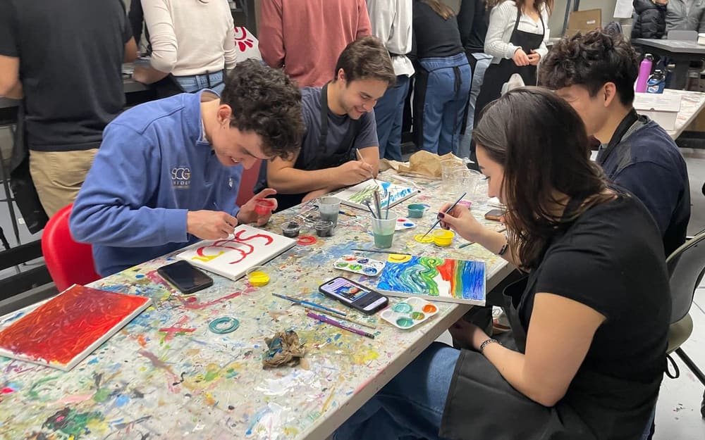 Members painting small canvases at a paint-spattered table, one in an SCG sweatshirt painting the SCG letters.