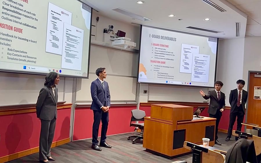 Four students in business attire presenting in a lecture room, one gesturing toward a screen showing slides titled E-Board Deliverables and Transition Guide.