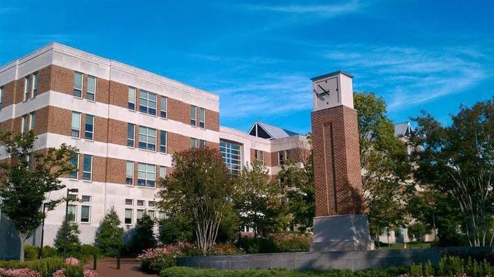 The Robert H. Smith School of Business building, brick and white stone, beside a brick clock tower, with trees and flower beds in front under a blue sky.