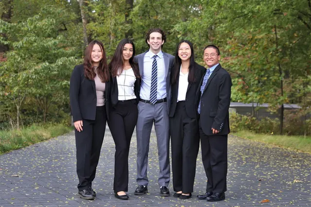 Five members in business attire standing together on a paved path in front of green trees.