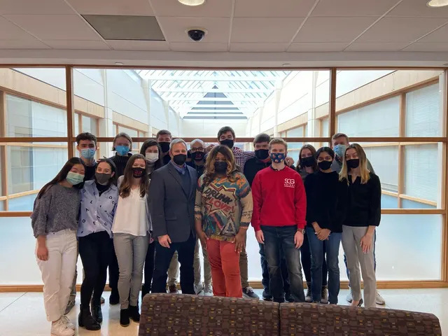 Members in casual clothes, most wearing face masks, gathered for a group photo in a bright hallway; one wears a red SCG sweatshirt.