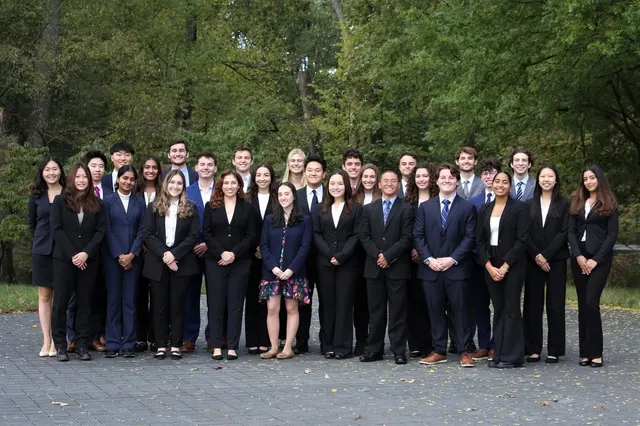 A large group of SCG members in business attire standing in rows on a paved area in front of trees.