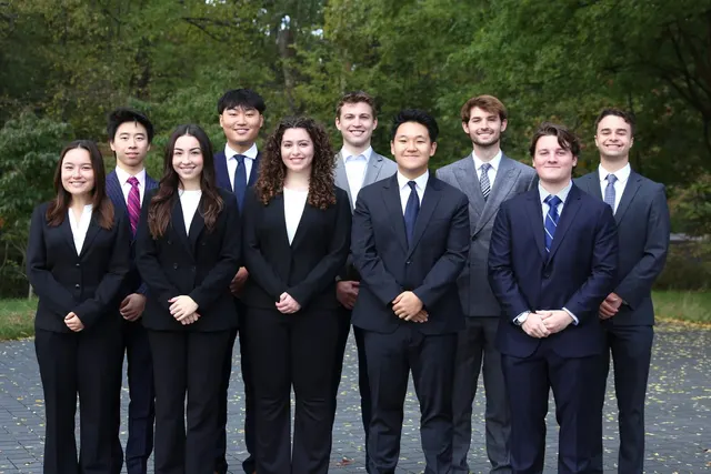SCG members in business attire standing in two rows on a paved area in front of tall trees.