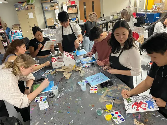 Members in aprons painting small canvases together at a long, paint-spattered art studio table.