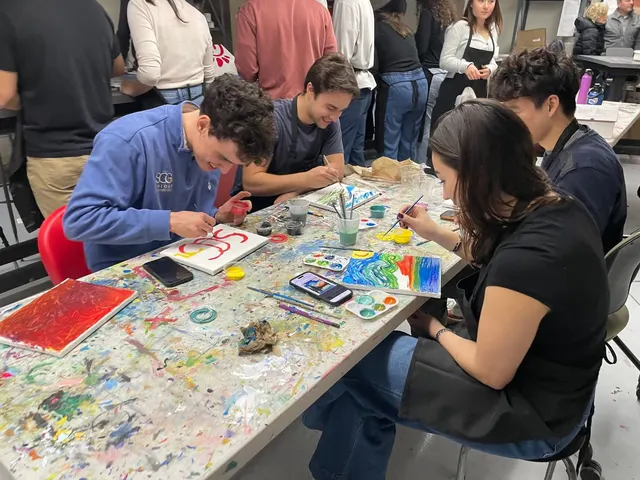 Members painting small canvases at a paint-spattered table, one in an SCG sweatshirt painting the SCG letters.
