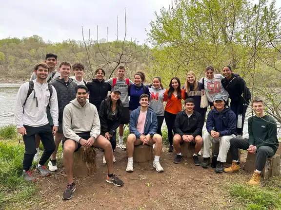 Members in casual clothes gathered by a river in spring, some sitting on tree stumps.