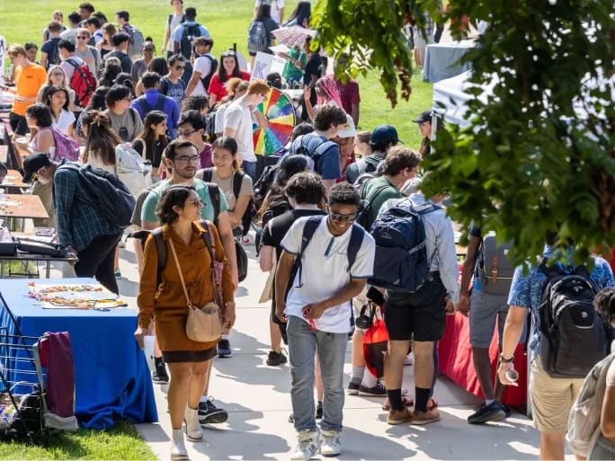 A crowd of students walking along a campus path past tables and booths at an outdoor fair on a sunny day.
