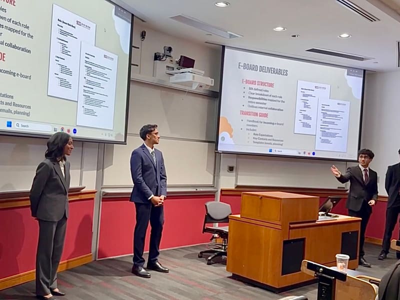 Five students in business attire presenting in a lecture room in front of two screens showing slides titled E-Board Deliverables and Transition Guide.