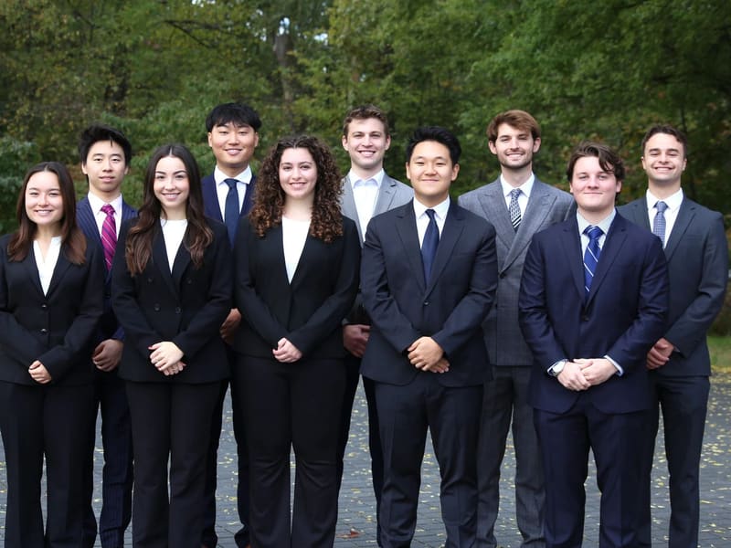SCG members in business attire standing in two rows on a paved area in front of tall trees.
